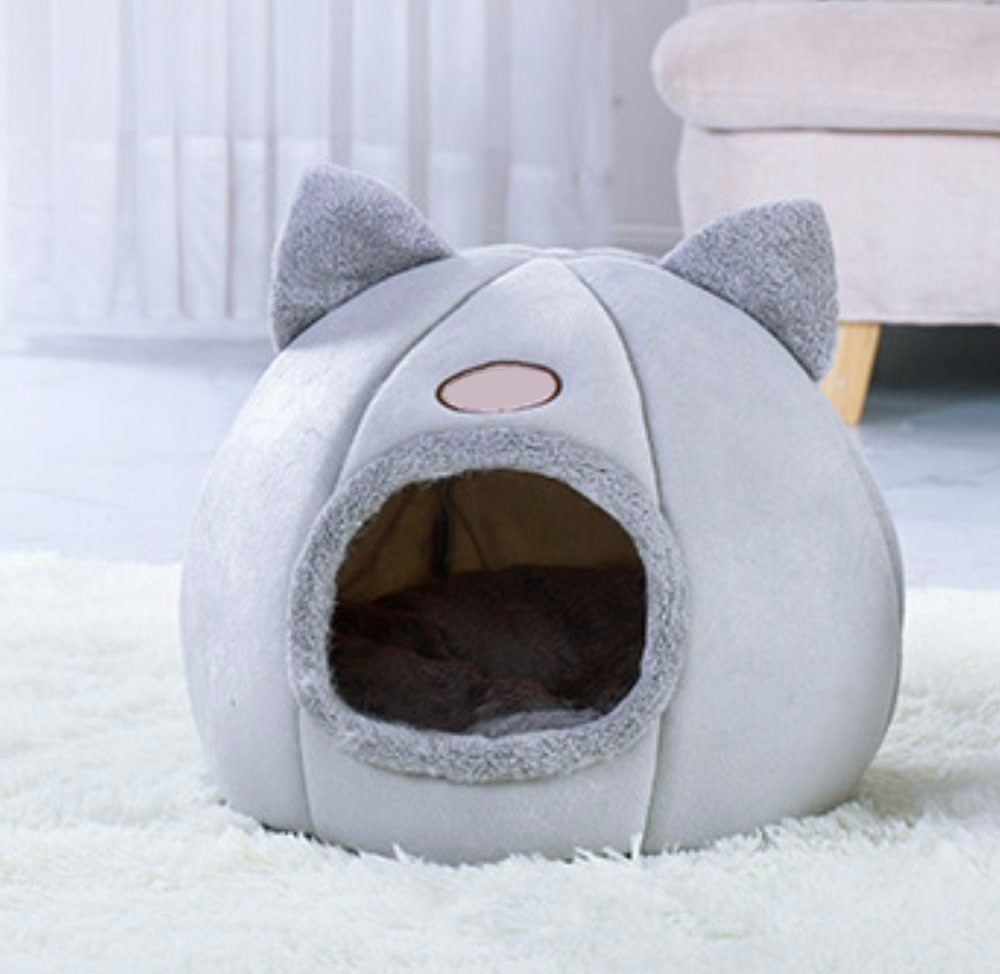 거실 러그 위에 놓인 회색 고양이 하우스, 둥근 몸통 위에 귀 두 개가 서 있고 앞쪽에 털 테두리를 두른 둥근 입구가 보인다